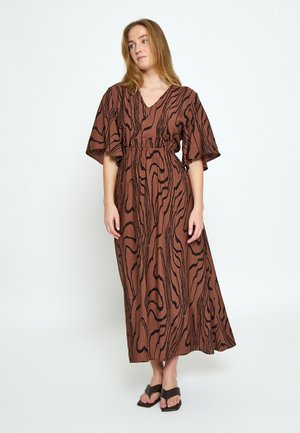Frau trägt ein braunes Maxikleid mit schwarzen abstrakten wellenförmigen Streifen und weiten kurzen Ärmeln, steht vor weißem Hintergrund.