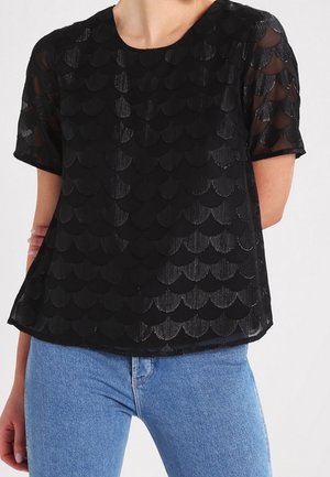 Kobieta ma na sobie czarny top z krótkim rękawem z falistym wzorem i przezroczystymi rękawami, połączony z jasnoniebieskimi jeansami z wysokim stanem.