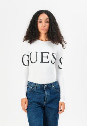 White long-sleeve top featuring large black “GUESS” lettering on the front, paired with blue high-waisted jeans. Smooth fabric texture.