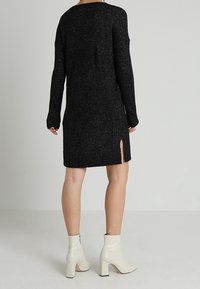 Zwarte gebreide jurk met lange mouwen, zijopeningen en een gestructureerde afwerking, gecombineerd met witte enkellaarsjes.