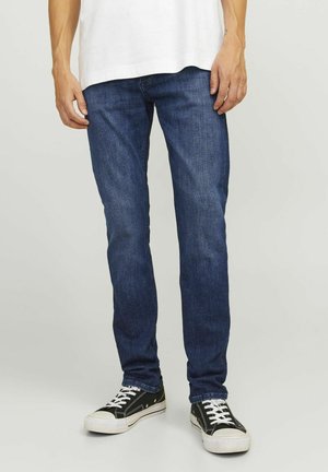 Jeans de denim azul oscuro con un corte slim, diseño de cinco bolsillos y costuras visibles. Combinados con zapatillas negras con acentos blancos.