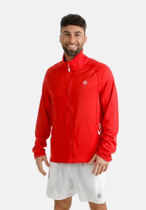 Ein lächelnder Mann trägt eine rote Jacke mit Reißverschluss und weiße Sportshorts, steht vor einem schlichten hellen Hintergrund.