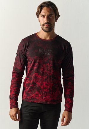 Hombre con cabello corto y oscuro y barba, vistiendo una camisa de manga larga con patrón rojo y negro y pantalones negros, de pie frente a un fondo claro y liso.