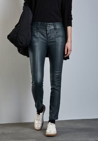 Pantalones de cuero ecológico ajustados de talle alto en verde oscuro, con dos bolsillos frontales, cierre de doble botón y textura suave. Combinados con zapatillas blancas.