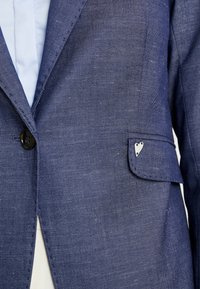 Blazer en denim bleu marine avec une finition texturée, doté d'un seul bouton noir et d'un petit ornement argenté en forme de cœur sur la poche poitrine.