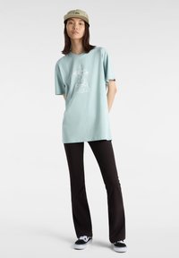 Camiseta de algodón azul claro con un diseño gráfico de dibujos animados en blanco, combinada con pantalones acampanados negros y zapatillas negras. Lleva una gorra verde.