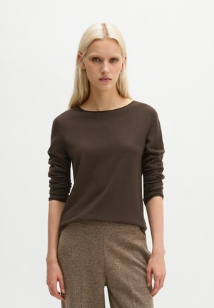 Femme aux longs cheveux blonds portant un haut à manches longues brun foncé et un pantalon texturé marron clair, debout devant un fond uni.