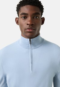 Lichtblauwe trui met kwartrits van zachte stof, voorzien van een hoge kraag en een smalle ritssluiting met een subtiel logodetail.