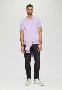 Jasnofioletowa koszulka polo z kołnierzykiem. Czarne jeansy slim-fit. Jasne trampki. Sweter zawiązany w pasie. Prosty krój, bez wzorów.