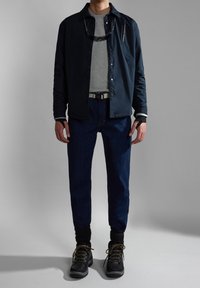 Chaqueta ligera de color azul marino sobre una camisa gris, jeans oscuros y botas de senderismo negras con cordones marrones. Presenta puños en capas y un cinturón.