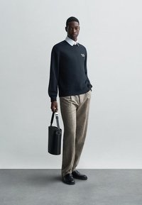 Čierna mikina s bielym logom, pod ňou svetlá košeľa s golierom, béžové vzorované nohavice, drží čiernu valcovú tašku, čierne topánky.