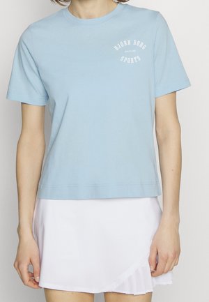 Person trägt hellblaues T-Shirt mit kurzen Ärmeln und dem Schriftzug „Björn Borg Sports“ sowie einen weißen plissierten Rock, steht vor einem einfarbigen Hintergrund.