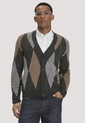 Mann mit klaren Brillen, weißem Hemd und einem dunkelgrauen, geknöpften Cardigan mit beige- und hellgrauem Argyle-Muster, stehend mit einer Hand in der Tasche.