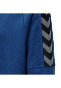 Tessuto blu con una cucitura a vista e un dettaglio a strisce chevron in bianco e nero lungo il bordo di una manica di un capo.