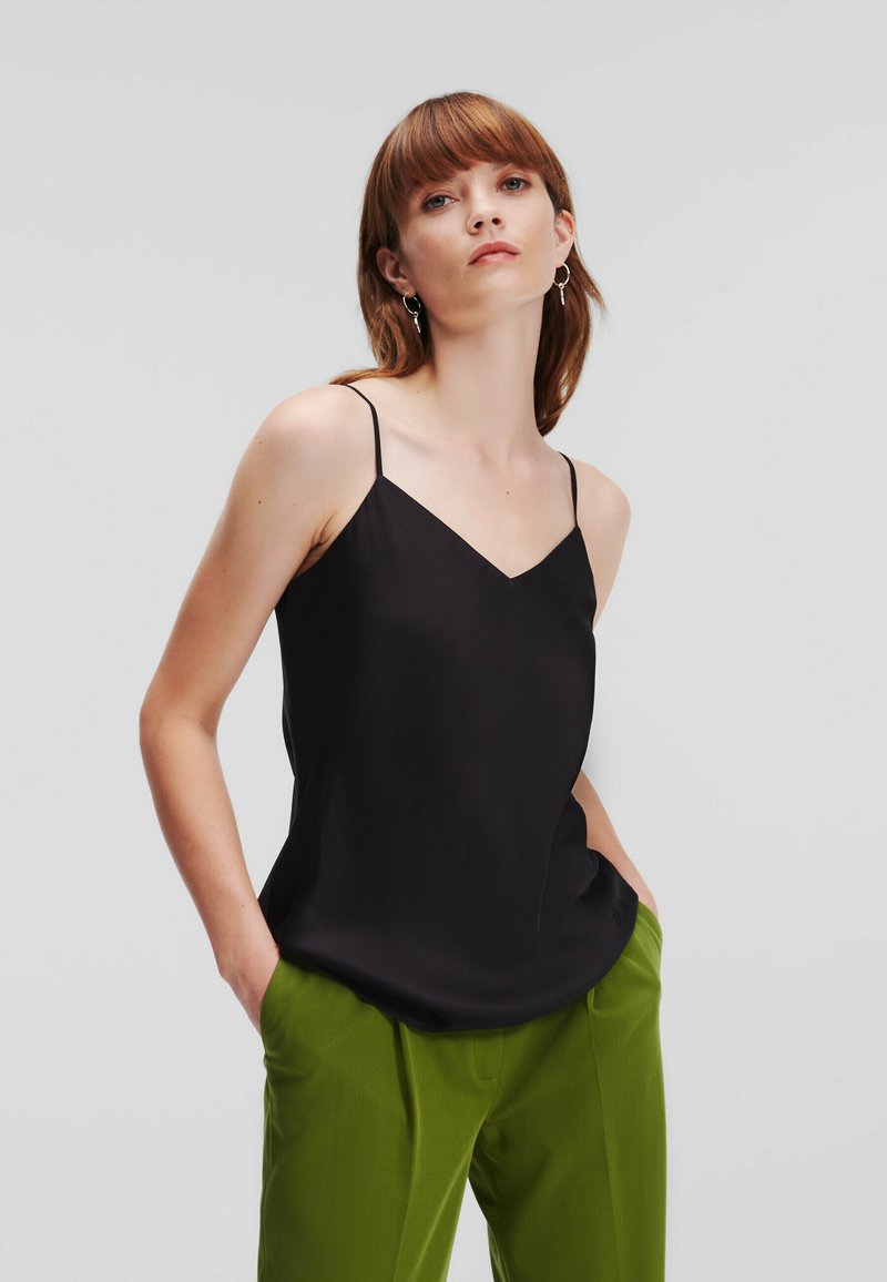 Camisola din satin negru cu bretele subțiri și design în V, asociată cu pantaloni verzi, evidențiind o textură netedă și o siluetă simplă.