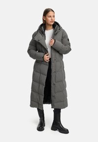 Grå pufferjakke med et langt design, hætte og knapforsyning, med en quiltet tekstur og store lommer. Båret over sorte bukser og støvler.