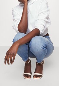 Biały krótki kaptur, jasnoniebieskie obcisłe dżinsy i beżowe szpilki z paskiem wokół kostki. Ręka spoczywa na kolanie, prezentując lakier na paznokciach.