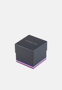 Scatola nera quadrata con coperchio, caratterizzata da un logo bianco "TOMMY HILFIGER" sulla parte superiore e una striscia di accento rossa, bianca e blu sulla base.