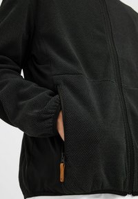 Crna jakna s teksturiranim mrežastim dizajnom, glatkim bočnim panelima, zatvaračem i malim kožnim povlačnim jezičkom na zatvaraču.