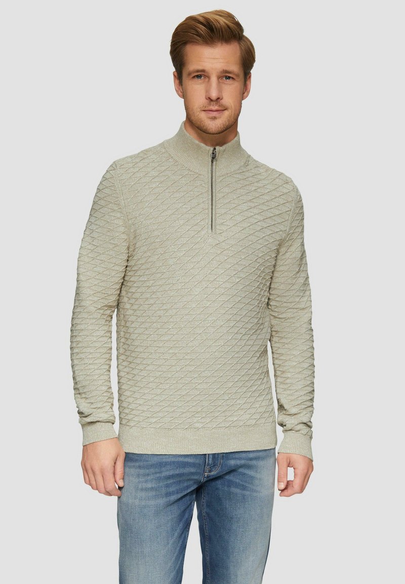 Hellbeige Zip-Up-Pullover mit Diamantstruktur, gerippten Bündchen und Saum, aus einem weichen Strickmaterial und kombiniert mit blauen Jeans.