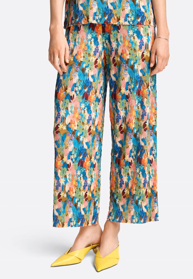 Frau in bunten, floral gemusterten Weitenhosen und passendem Oberteil mit gelben spitz zulaufenden Slip-On Schuhen vor weißem Hintergrund.