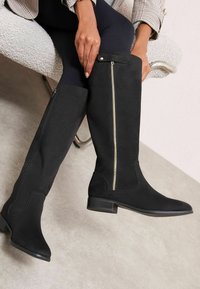 Egy személy ül egy texturált széken, fekete térdig érő csizmában, oldalsó cipzárral, sötét leggingsben és kockás kabátban.
