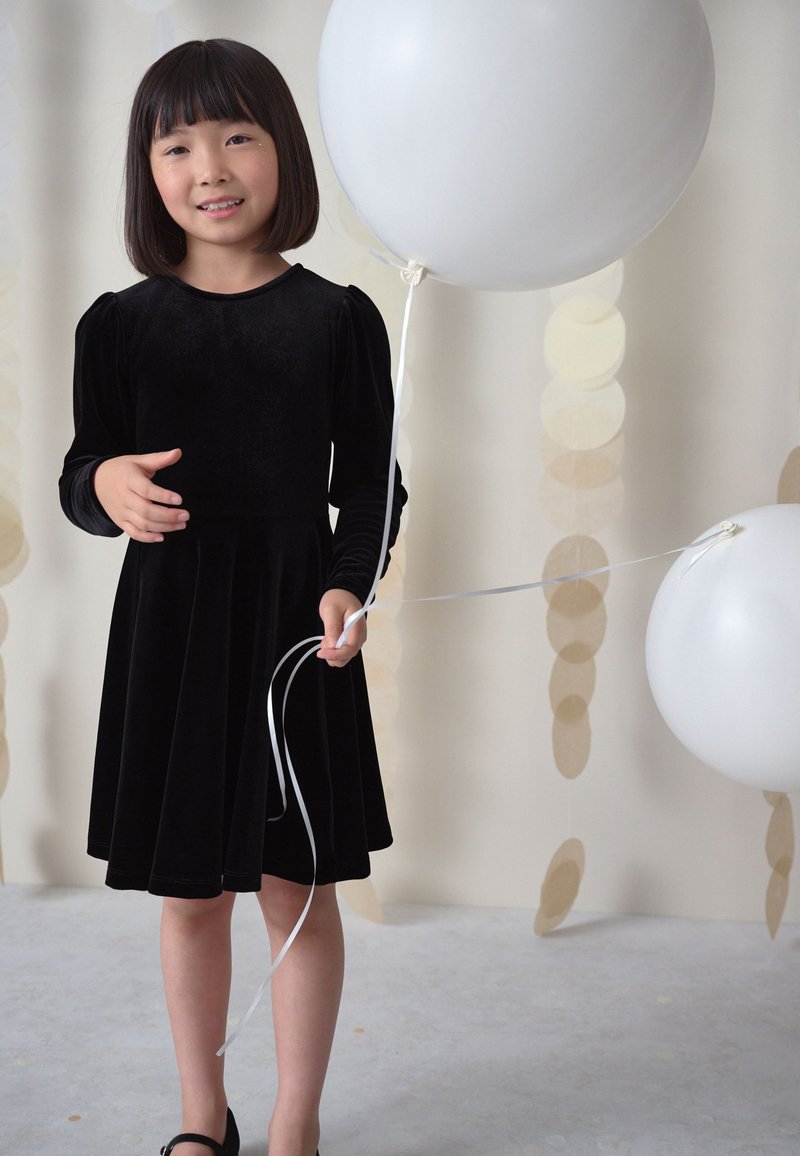 Robe en velours noir à manches longues et jupe évasée ; tenant des ballons blancs avec de longues ficelles blanches devant un fond clair.