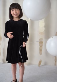 Schwarzes Samtkleid mit langen Ärmeln und einem ausgestellten Rock; hält weiße Luftballons mit langen, weißen Schnüren vor einem hellen Hintergrund.
