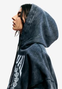 Ciemnoszara bluza z kapturem z dużym logo z przodu, wykonana z teksturowanej tkaniny, z kapturem ze ściągaczem i luźnym krojem, widoczna z profilu.