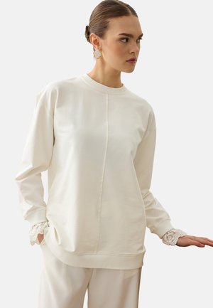 Femme portant un haut crème à manches longues avec un détail en dentelle aux poignets et un pantalon ample assorti, regardant sur le côté devant un fond uni.