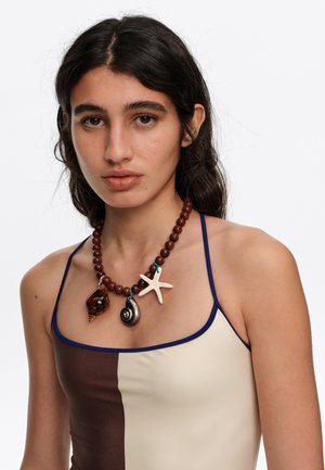 Joven mujer con cabello oscuro que lleva una camiseta de tirantes con bloques de color marrón y crema y un collar de cuentas de madera con colgantes de conchas marinas y estrellas de mar.