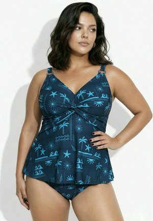 Frau mit dunklem, welligem Haar, die einen dunkelblauen Tankini mit hellblauen tropischen Mustern trägt und mit einer Hand auf der Hüfte steht.