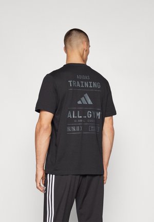 Man draagt een zwart Adidas trainingsshirt met tekst en logo op de achterkant, gecombineerd met zwarte Adidas broek met witte strepen.
