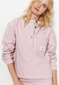 Chemise à rayures en coton doux rose pâle et blanc, avec un col montant, une poche avant et des détails de couture visibles.