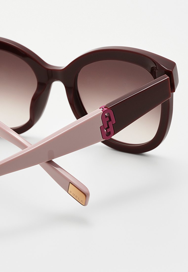 Lunettes de soleil avec de grandes montures bourgogne foncé, des verres dégradés et des branches rose clair ornées d'un logo violet et d'accents métalliques.