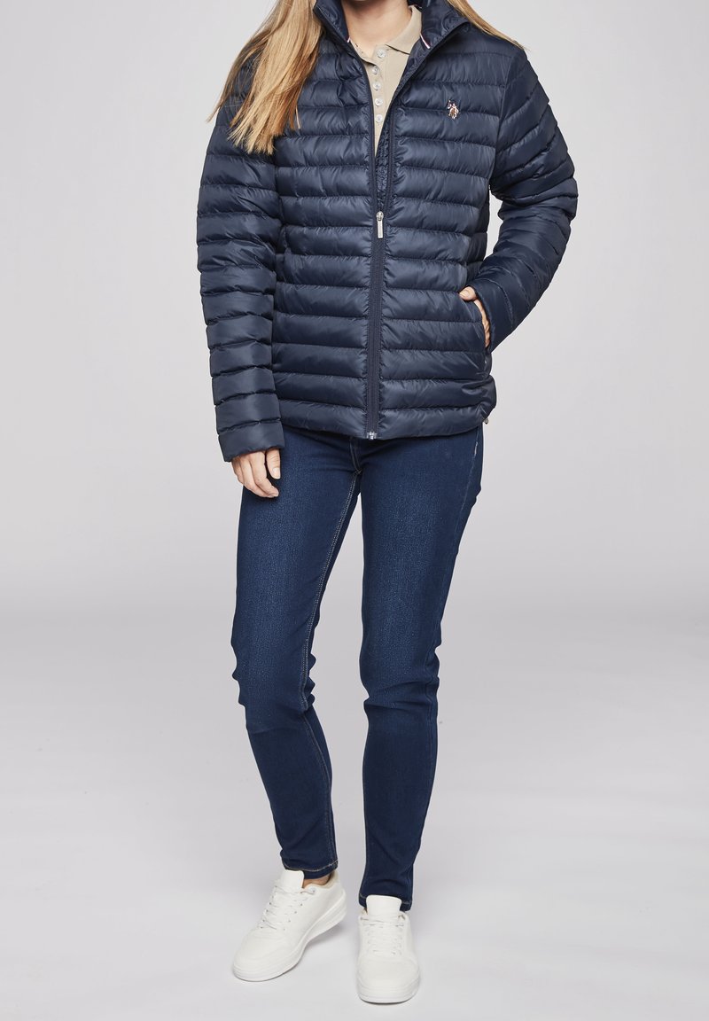 Persoon die een marineblauwe doorgestikte jas, beige poloshirt, donkerblauwe spijkerbroek en witte sneakers draagt, staand tegen een effen achtergrond.