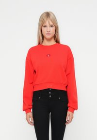 Sweatshirt rouge court en matériau doux avec des manches longues. Présente un petit pin en forme de cœur avec du texte, associé à un jean noir taille haute.