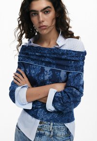 Blau gemusterte Bluse mit gerippter Textur, die über einem hellblauen Hemd mit Kragen getragen wird. Verfügt über lange Ärmel mit geknöpften Manschetten. Jeans sind sichtbar.