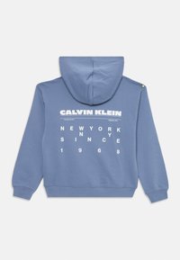 Kootiväega särk helesinisest kangast, millel on valge tekst: "CALVIN KLEIN," "NEW YORK" ja "SINCE 1968," ning kahe nööbiga õlal.