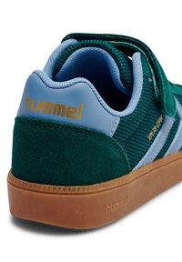 Zielony zamszowy i siateczkowy trampek z wyściełanym niebieskim kołnierzem, brązową gumową podeszwą i złotym logo "hummel" na pięcie, z paskowanymi akcentami.