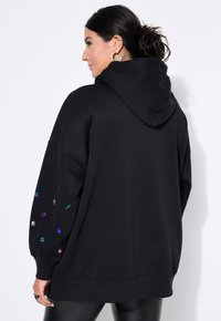 Černá oversized mikina vyrobená z měkké tkaniny, se kapucí a zdobená barevnými akcenty ve tvaru drahokamů na rukávech.
