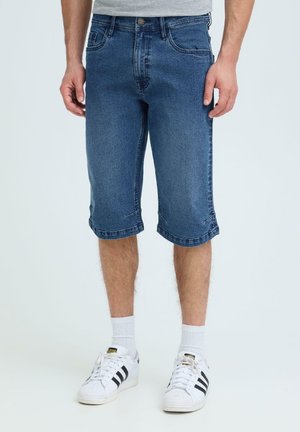 Uomo che indossa pantaloncini di denim blu fino al ginocchio, calzini bianchi e scarpe da ginnastica Adidas bianche con strisce nere, in piedi davanti a uno sfondo semplice.