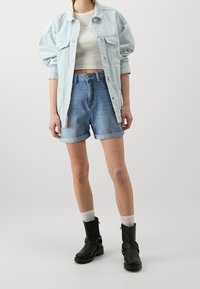 Chaqueta de mezclilla azul claro sobre una camiseta corta blanca, combinada con shorts azules remangados y botas negras hasta el tobillo. Los accesorios incluyen un collar de cadena.