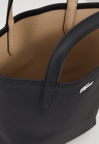 Sac fourre-tout noir avec un extérieur texturé, doté d'une doublure beige. Comprend deux anses robustes et un petit détail de logo métallique.