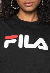 T-shirt noir arborant un logo FILA blanc audacieux avec une touche de rouge. Le tissu est lisse et le t-shirt possède un col rond.