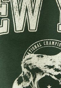 Tmavě zelené bavlněné tričko s velkým opotřebovaným bílým textem a grafikou vlka, s kruhovým textem rámujícím design.