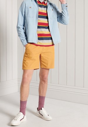 Helles blaues Denim-Jackett, gestreiftes Polo in Grau, Marineblau und Rot, gelbe Shorts, kastanienbraune Socken und weiße Sneakers mit braunen Akzenten.