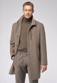 Cappotto in lana marrone con colletto, bottoni frontali e tasche laterali. Indossato sopra un dolcevita coordinato e pantaloni grigi su misura. Tessuto liscio.