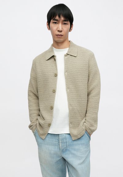 Svetlo béžový textúrovaný kardigán s golierom, zapínanie na gombíky a dlhé rukávy, nosený cez bielu košeľu a svetlé rifľové nohavice.