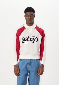 Vit och röd stickad polo tröja med långa ärmar, med en svart "obey"-logotyp i mitten och ribbade cuffs och nederkant. Bärs med blå jeans.